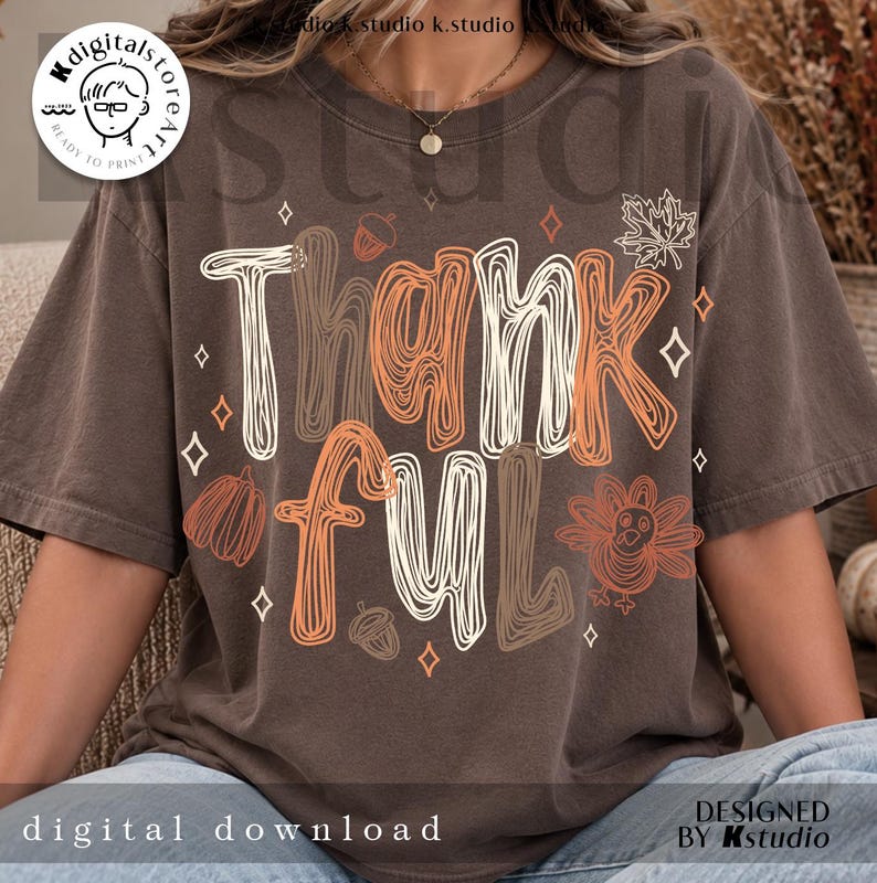 K&ouml;nnte beinhalten: Ein dunkelbraunes Kurzarm-T-Shirt mit dem Wort Thankful in einer stilisierten, mehrfarbigen Linienkunst-Schriftart. Das Design enth&auml;lt orangefarbene und wei&szlig;e Umrisse eines Truthahns, K&uuml;rbissen, Eicheln und kleinen vierzackigen Sternen.