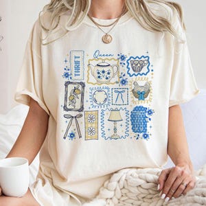May include: Cream-colored t-shirt featuring a collage of vintage-style illustrations in blue and yellow. The design includes a teapot, teacup, lemon, and a hen in a basket. The word "Thrift" is printed vertically.