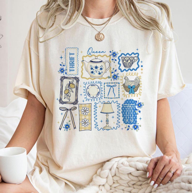 May include: Cream-colored t-shirt featuring a collage of vintage-style illustrations in blue and yellow. The design includes a teapot, teacup, lemon, and a hen in a basket. The word "Thrift" is printed vertically.