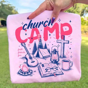K&ouml;nnte beinhalten: Ein hellrosa Sweatshirt mit einem grafischen Design mit den Worten "church CAMP" in Rosa und Blau. Das Design umfasst eine Gitarre, ein Zelt, eine Laterne, ein Kreuz, ein Lagerfeuer, ein Buch und eine Kaffeetasse. Die Worte "faith friends fun" sind ebenfalls enthalten.