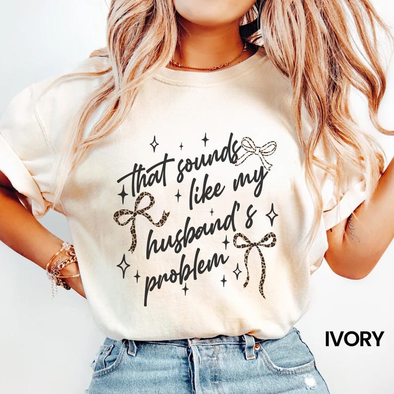 May include: Ivory-colored t-shirt with the phrase "That sounds like my husband's problem" in black script. The design includes bow accents with leopard print and star details. The word "IVORY" is printed at the bottom.