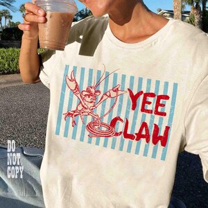 May include: Cream-colored t-shirt featuring a red cartoon lobster wearing a cowboy hat and holding a lasso, set against blue and white vertical stripes. The words "YEE CLAW" are printed in red. The person is holding a clear plastic cup with a brown beverage.