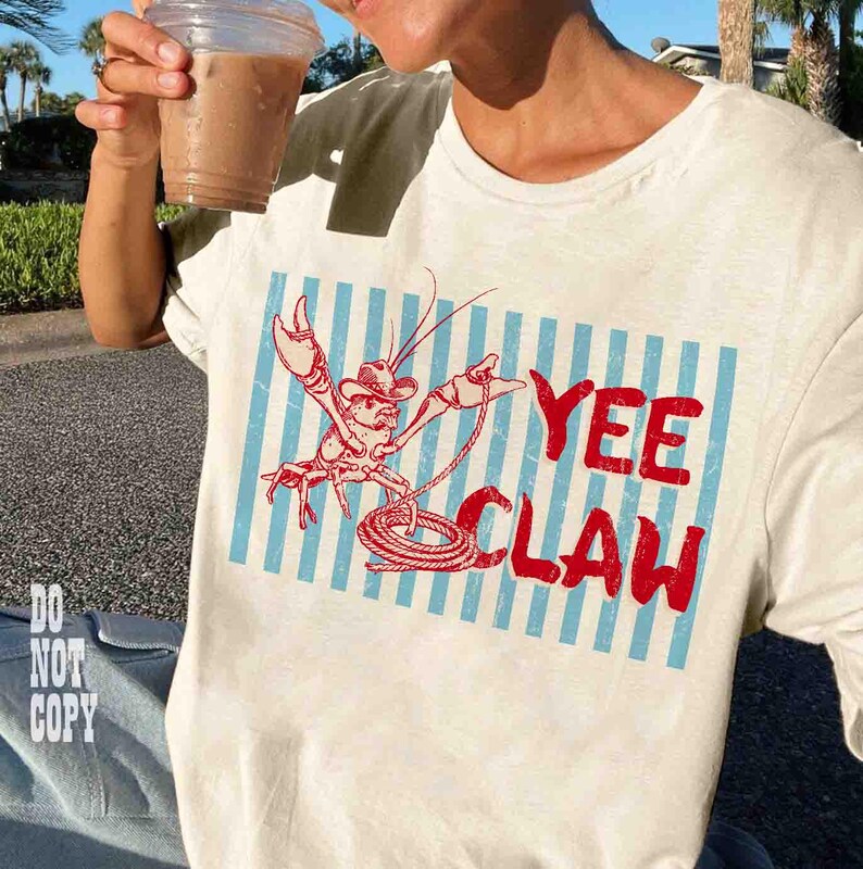 May include: Cream-colored t-shirt featuring a red cartoon lobster wearing a cowboy hat and holding a lasso, set against blue and white vertical stripes. The words "YEE CLAW" are printed in red. The person is holding a clear plastic cup with a brown beverage.