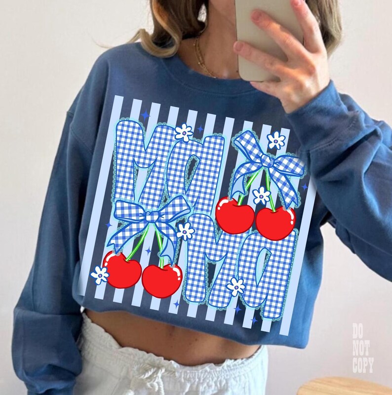 May include: A blue sweatshirt with the word "MAMA" in a gingham pattern, decorated with red cherries, blue bows, and white flowers. The sweatshirt has a striped design and long sleeves.