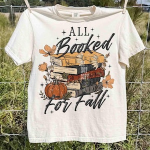 May include: Cream-colored t-shirt with the text "ALL Booked for Fall" in black script. The graphic features a stack of books, a pumpkin, and floral accents in orange and brown. The shirt is hanging on a clothesline.