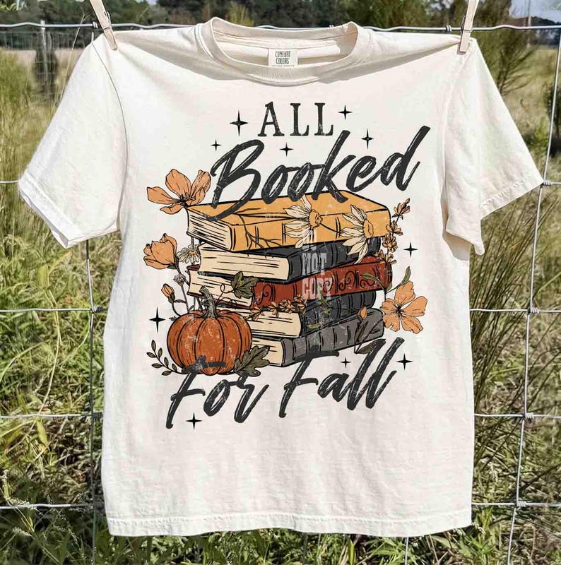 May include: Cream-colored t-shirt with the text "ALL Booked for Fall" in black script. The graphic features a stack of books, a pumpkin, and floral accents in orange and brown. The shirt is hanging on a clothesline.