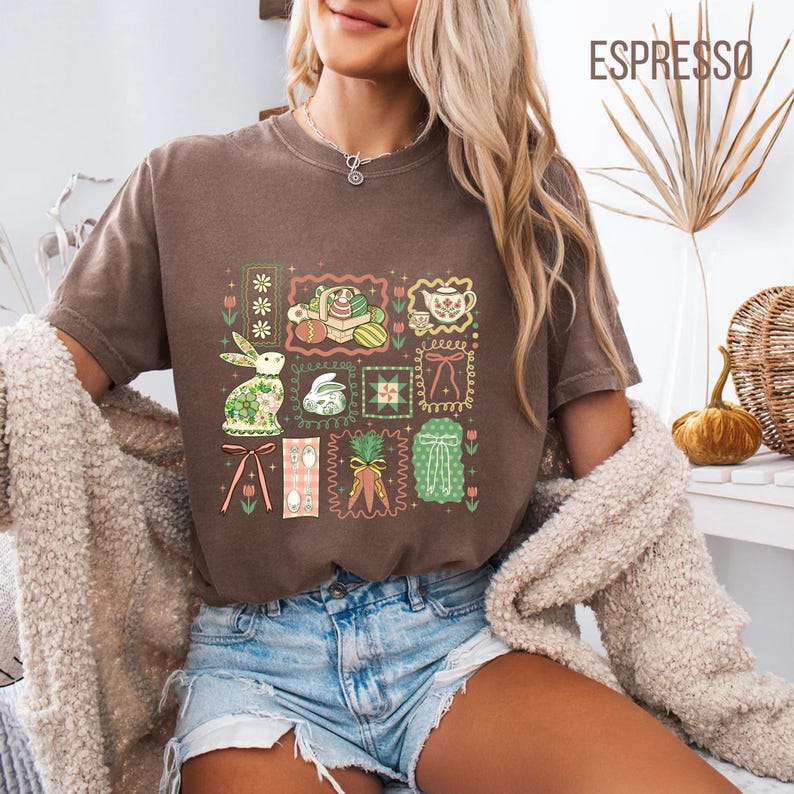 May include: Espresso-colored t-shirt featuring a colorful Easter-themed graphic. The design includes a bunny, eggs, carrots, and other spring elements within decorative frames. The shirt has short sleeves and a relaxed fit, perfect for casual wear.