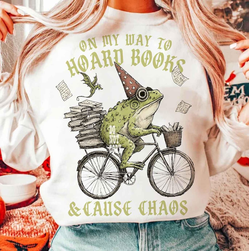 May include: White sweatshirt featuring a frog on a bicycle, wearing a wizard hat, and carrying books. The text reads "ON MY WAY TO HOARD BOOKS & CAUSE CHAOS". A dragon and floating papers are also depicted in the illustration.