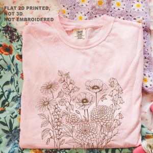May include: A light pink t-shirt featuring a detailed brown floral design. The design includes various flowers such as daisies, poppies, and bellflowers. The text "FLAT 2D PRINTED, NOT 3D, NOT EMBROIDERED" is printed at the top left.