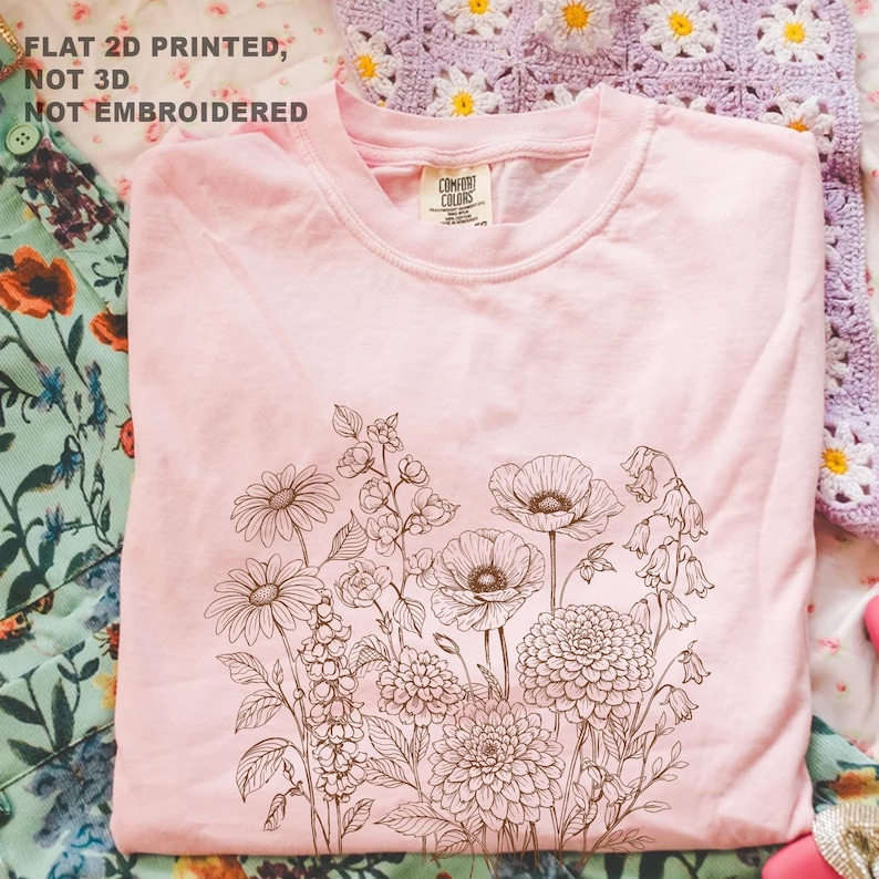 May include: A light pink t-shirt featuring a detailed brown floral design. The design includes various flowers such as daisies, poppies, and bellflowers. The text "FLAT 2D PRINTED, NOT 3D, NOT EMBROIDERED" is printed at the top left.
