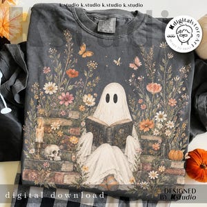 Op de afbeelding: Een antracietgrijs t-shirt met een afbeelding van een witte geest die een boek leest terwijl deze op een stapel oude boeken zit. Het ontwerp bevat kleurrijke wilde bloemen, vlinders, een brandende kaars en een schedel in een illustratie.