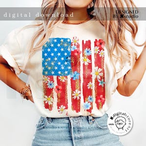 Puede incluir: Una camiseta blanca con un dise&ntilde;o de bandera estadounidense desgastada con rayas y estrellas rojas, blancas y azules. La bandera est&aacute; decorada con flores blancas y azules. El texto "DESIGNED BY Kstudio" est&aacute; en la esquina superior derecha de la imagen.