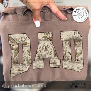 May include: A mauve-colored sweatshirt featuring the word "DAD" in a distressed, block letter design. The letters are filled with a detailed illustration of ducks, reeds, and water. The words "digital download" and "designed by k.studio" are also visible.