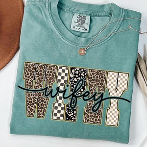 May include: A teal Comfort Colors t-shirt featuring the word "Wifey" in a decorative font. The letters are a mix of leopard print, black and white checkered, and tan checkered patterns. A gold necklace with a pendant is on the shirt.