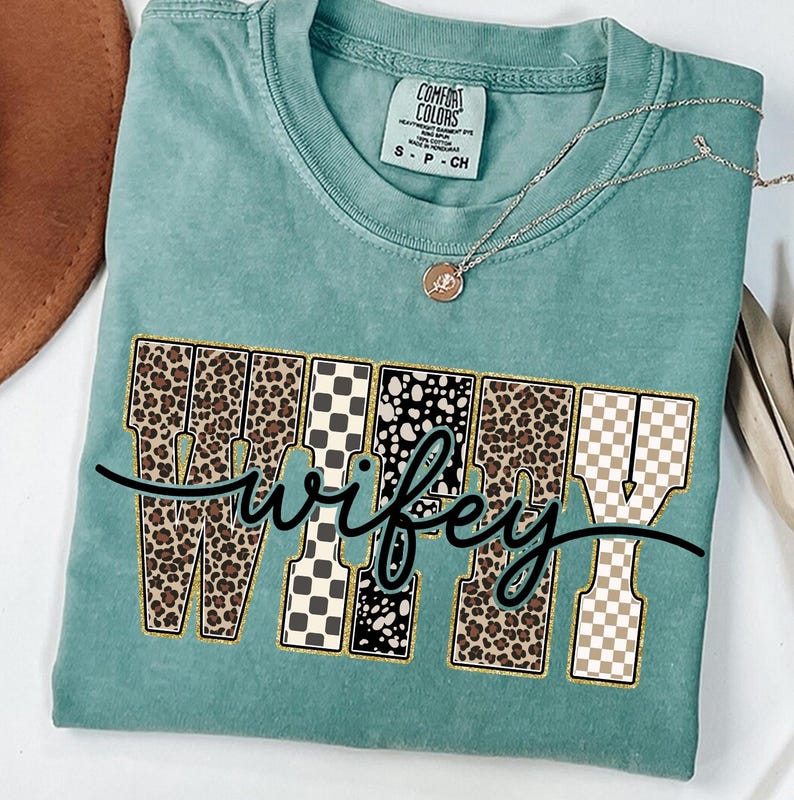 May include: A teal Comfort Colors t-shirt featuring the word "Wifey" in a decorative font. The letters are a mix of leopard print, black and white checkered, and tan checkered patterns. A gold necklace with a pendant is on the shirt.