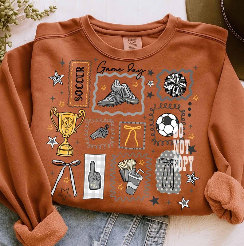 May include: Rust-coloured sweatshirt with a soccer theme. The design includes a trophy, soccer ball, cleats, pom-pom, and the words "Game Day" and "Soccer." The phrase "Do Not Copy" is also visible. The sweatshirt has a fleece lining.