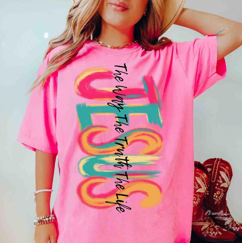 May include: A bright pink t-shirt with the word "JESUS" in large, colorful brushstrokes. The text "The Way The Truth The Life" is printed vertically on the left side of the word "JESUS". The shirt is paired with red cowboy boots.