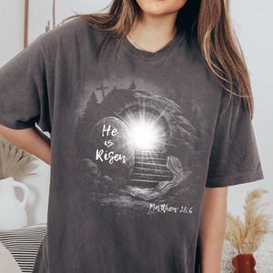 May include: Dark gray t-shirt featuring a graphic of an open tomb with a bright light emanating from within. The text "He is Risen" is displayed on a stone, with "Matthew 28:6" below the image.
