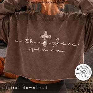 Op de afbeelding: Een bruin, langmouwige, cropped sweatshirt met een witte afbeelding van een krabbelkruis en de tekst "with Jesus you can." Het kledingstuk heeft een ronde hals, lange mouwen en een zoom met een rafelige rand, met een vervaagde afwerking.