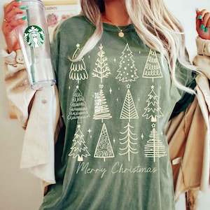 May include: Olive green t-shirt featuring a collection of illustrated Christmas trees in various styles. The words "Merry Christmas" are printed in a cursive font at the bottom of the design. The shirt is made of a soft material.