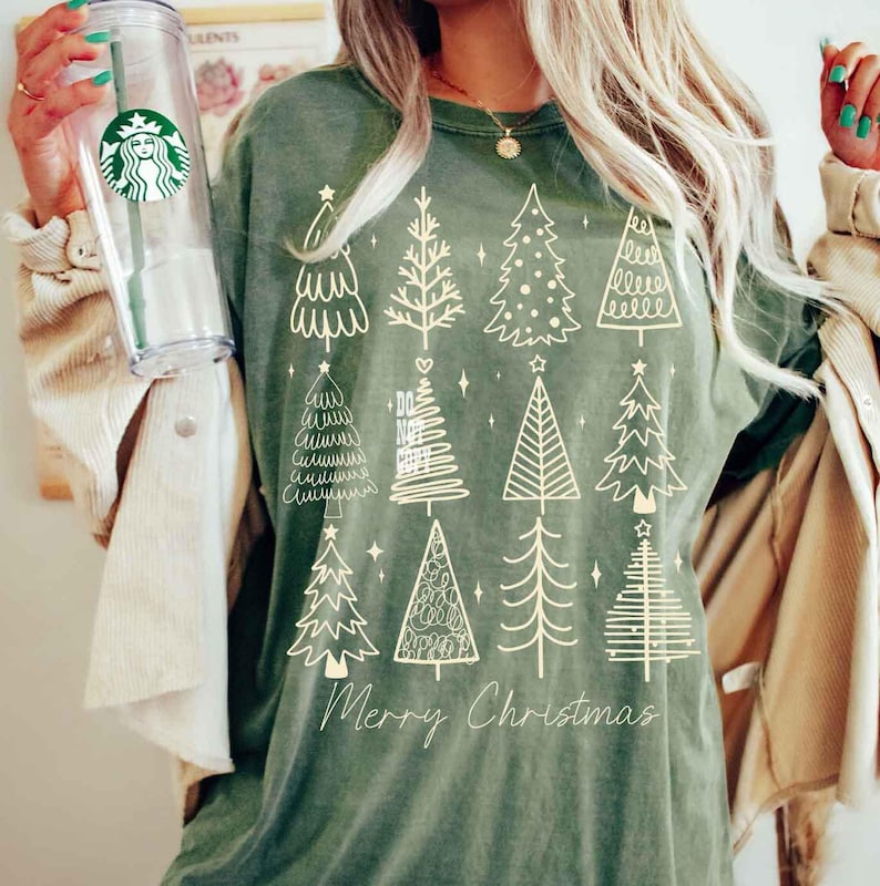 May include: Olive green t-shirt featuring a collection of illustrated Christmas trees in various styles. The words "Merry Christmas" are printed in a cursive font at the bottom of the design. The shirt is made of a soft material.