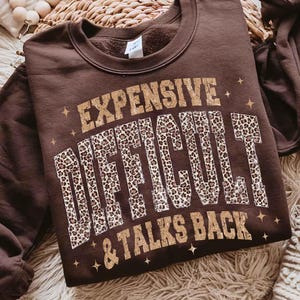 May include: A brown crew neck sweatshirt featuring the text "EXPENSIVE DIFFICULT & TALKS BACK" in gold and leopard print. The sweatshirt is made of a soft material. The design includes star accents.