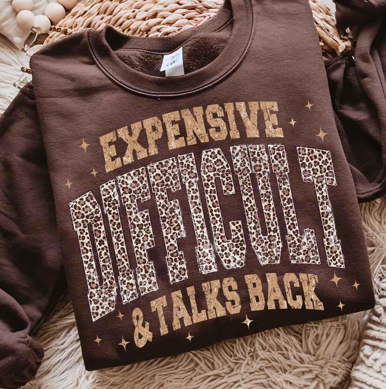 May include: A brown crew neck sweatshirt featuring the text "EXPENSIVE DIFFICULT & TALKS BACK" in gold and leopard print. The sweatshirt is made of a soft material. The design includes star accents.