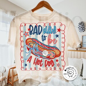 May include: Cream-colored t-shirt with a graphic of a hot dog and the text "BAD day to be A Hot Dog." The design includes red, blue, and pink colors, with stars and a wavy border. The shirt is hanging on a wooden hanger.