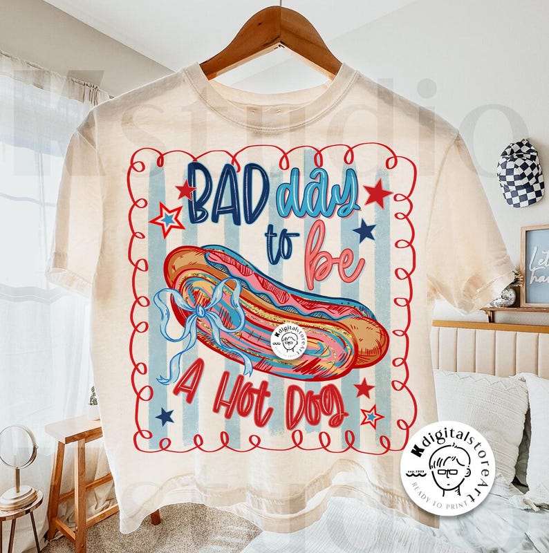 May include: Cream-colored t-shirt with a graphic of a hot dog and the text "BAD day to be A Hot Dog." The design includes red, blue, and pink colors, with stars and a wavy border. The shirt is hanging on a wooden hanger.