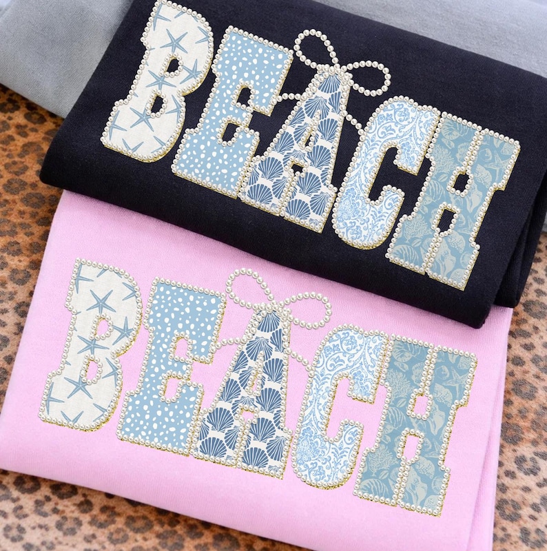 May include: Two folded t-shirts, one black and one pink, each featuring the word "BEACH" in large, decorative letters. The letters are decorated with light blue patterns and faux pearls. The "A" has a pearl bow.