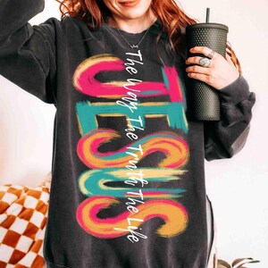 May include: Dark gray sweatshirt with the word "JESUS" in large, colorful, brushstroke letters. The text "The Way The Truth The Life" is written vertically along the left side of the word "JESUS". The sweatshirt is a crew neck style.