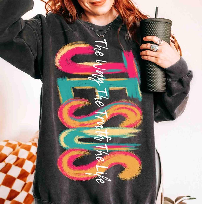 May include: Dark gray sweatshirt with the word "JESUS" in large, colorful, brushstroke letters. The text "The Way The Truth The Life" is written vertically along the left side of the word "JESUS". The sweatshirt is a crew neck style.