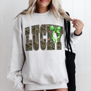 May include: A light gray sweatshirt with the word "LUCKY" in a camouflage pattern with a green ribbon. The sweatshirt is a casual style, suitable for everyday wear.