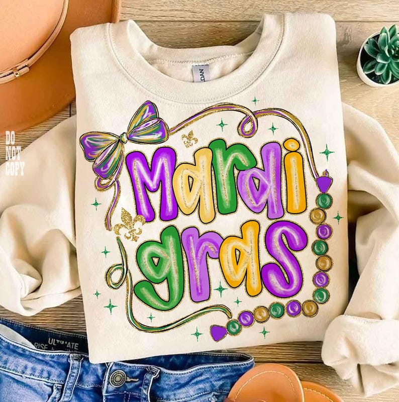 May include: Cream-colored sweatshirt with a colorful "Mardi Gras" design. The design features the text in green, purple, and yellow, with a bow, fleur-de-lis, and bead accents. The sweatshirt is folded on a wooden surface.