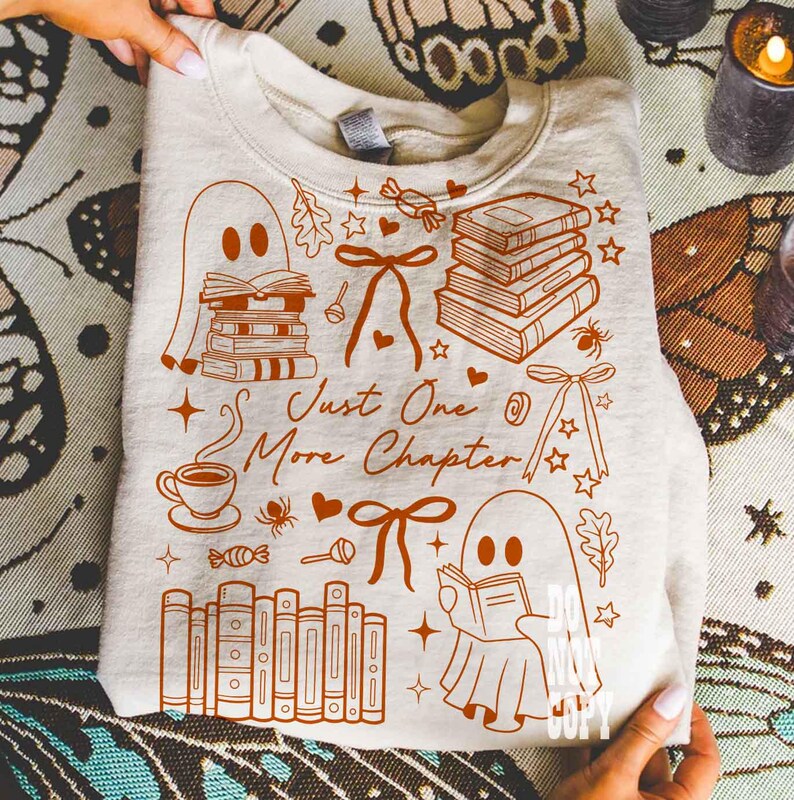 May include: A beige sweatshirt with a brown graphic design featuring ghosts reading books, stacks of books, bows, stars, and the text "Just One More Chapter." The design includes a coffee cup, candy, and a spider.