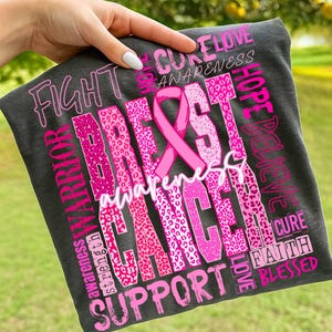 Op de afbeelding: Donkergrijs t-shirt met een roze borstkanker bewustzijds lint en tekst. De woorden "Fight", "Warrior", "Support", "Awareness", "Breast Cancer", "Hope", "Believe", "Cure", "Love", "Faith" en "Blessed" staan op het shirt.