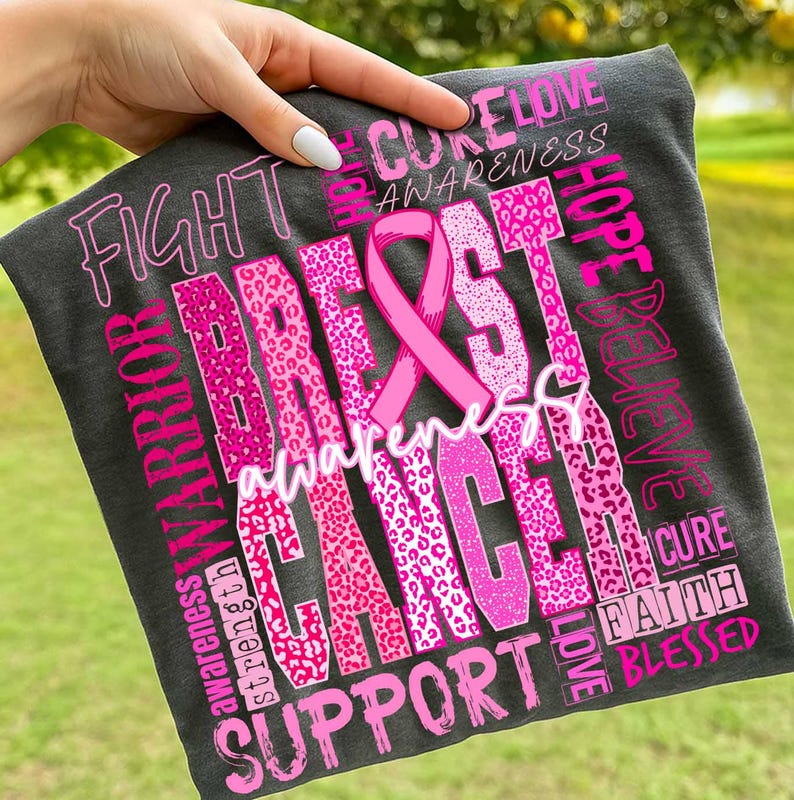 Op de afbeelding: Donkergrijs t-shirt met een roze borstkanker bewustzijds lint en tekst. De woorden "Fight", "Warrior", "Support", "Awareness", "Breast Cancer", "Hope", "Believe", "Cure", "Love", "Faith" en "Blessed" staan op het shirt.