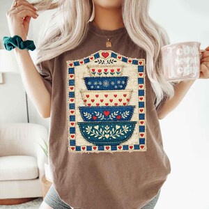 Pu&ograve; includere: T-shirt marrone con un design a ciotole impilate con cuori rossi e motivi floreali blu. Il design &egrave; incorniciato da un bordo blu e accenti di cuori rossi. La maglietta &egrave; in stile casual.