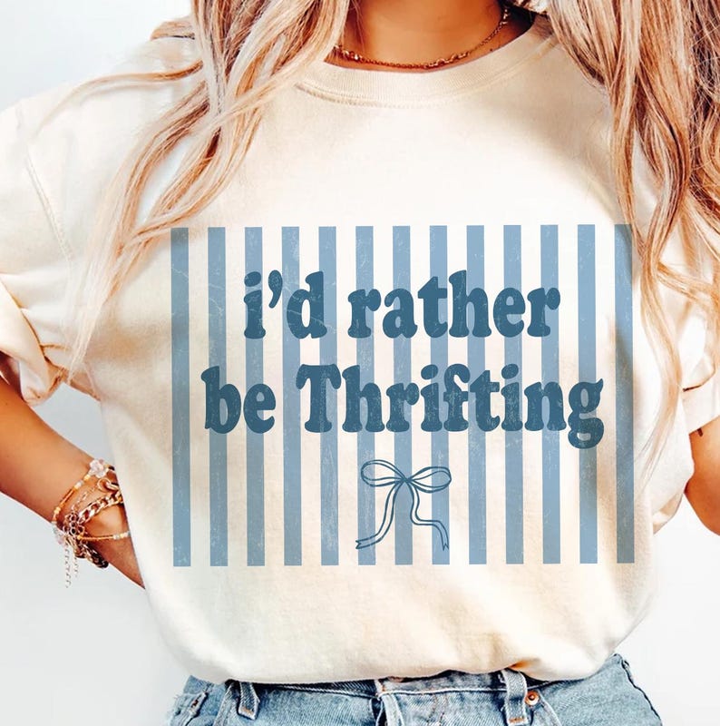 May include: Cream-colored t-shirt with the text "I'd rather be Thrifting" in blue. The text is set against a backdrop of vertical blue stripes and a bow graphic. The shirt is a casual, everyday wear item.