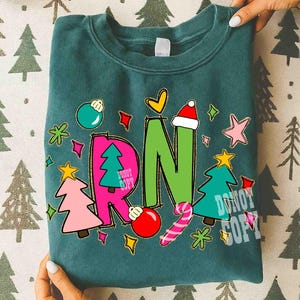 May include: Teal sweatshirt with a colorful Christmas design. The design includes the letters "RN" in green and pink, decorated with Christmas ornaments, trees, and a Santa hat. The words "DO NOT COPY" are also visible.