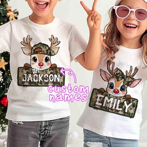 May include: Two white t-shirts with a cartoon reindeer design. The reindeer wears a camouflage hat and holds a sign with a custom name. One shirt says "JACKSON" and the other says "EMILY".