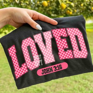 May include: Dark gray t-shirt with the word "LOVED" in large, pink, checkered letters. Below the word "LOVED" is the text "JOHN 3:16" in a pink rectangular shape. The shirt is held up by a hand.
