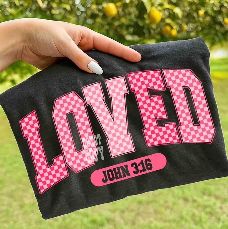 May include: Dark gray t-shirt with the word "LOVED" in large, pink, checkered letters. Below the word "LOVED" is the text "JOHN 3:16" in a pink rectangular shape. The shirt is held up by a hand.