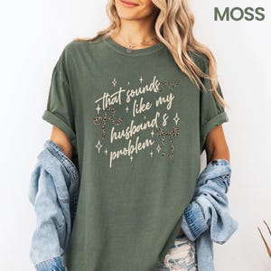 May include: Moss green t-shirt with rolled-up sleeves. The shirt features the text "That sounds like my husband's problem" in a script font, with leopard print bow accents and star details. The word "MOSS" is in the upper right corner.