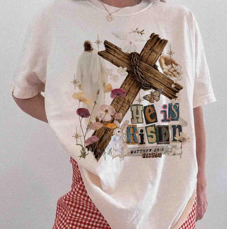 May include: Cream-colored t-shirt featuring a graphic design with a wooden cross, flowers, a rabbit, and the text "He is Risen" with "Matthew 28:6" below. The design incorporates pastel colors and a vintage aesthetic.