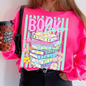 May include: A bright pink sweatshirt with the words "BOOK LOVER" in a playful font. The design features a stack of colorful books adorned with flowers. The sweatshirt also has vertical teal stripes.