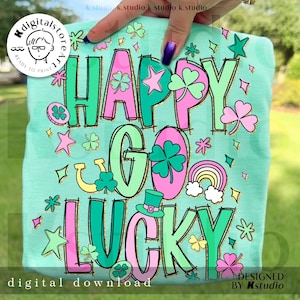 Op de afbeelding: Lichtgroen T-shirt met de tekst "HAPPY GO LUCKY" in roze en groen, versierd met klavers, sterren, een regenboog en een kabouterhoed. De woorden "digital download" en "designed by Kstudio" zijn ook zichtbaar.