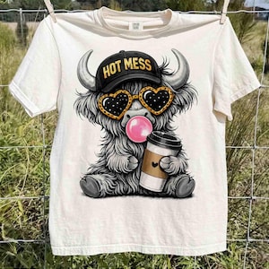 K&ouml;nnte beinhalten: Cremefarbenes T-Shirt mit einer Cartoon-Highland-Kuh, die eine herzf&ouml;rmige Sonnenbrille, eine schwarze Kappe mit dem Text "HOT MESS" tr&auml;gt und eine Kaugummiblase bl&auml;st. Die Kuh h&auml;lt eine Kaffeetasse mit einem Herz. Das Shirt h&auml;ngt an einer W&auml;scheleine.