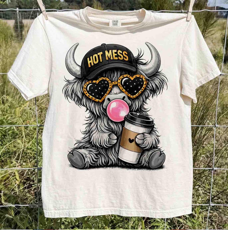 K&ouml;nnte beinhalten: Cremefarbenes T-Shirt mit einer Cartoon-Highland-Kuh, die eine herzf&ouml;rmige Sonnenbrille, eine schwarze Kappe mit dem Text "HOT MESS" tr&auml;gt und eine Kaugummiblase bl&auml;st. Die Kuh h&auml;lt eine Kaffeetasse mit einem Herz. Das Shirt h&auml;ngt an einer W&auml;scheleine.
