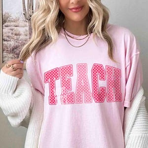 K&ouml;nnte beinhalten: Hellrosa T-Shirt mit dem Wort "TEACH" in einem rosa karierten Design. Das Shirt hat einen Rundhalsausschnitt und kurze &Auml;rmel. Der Stoff wirkt weich und bequem. Der Stil ist l&auml;ssig.